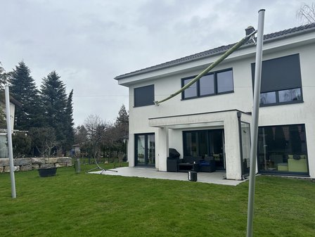 Einfamilienhaus mit Terrasse und Rasen, davor schräg gespannte grüne Stange zwischen Metallpfosten.
