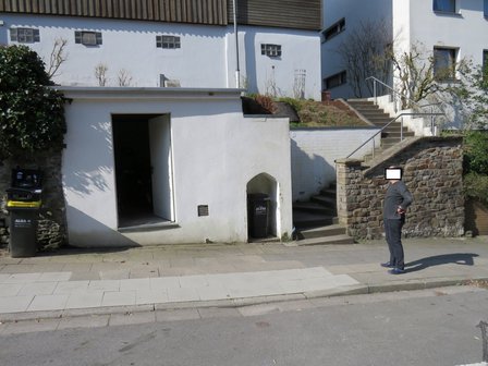 Mann steht vor weißem Gebäudesockel mit offener Tür, Treppe und Steinmauer.