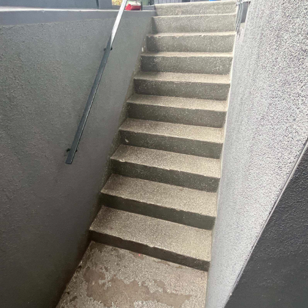 Graue Betontreppe mit Handlauf zwischen verputzten Wänden, unten feuchte Flecken auf dem Podest
