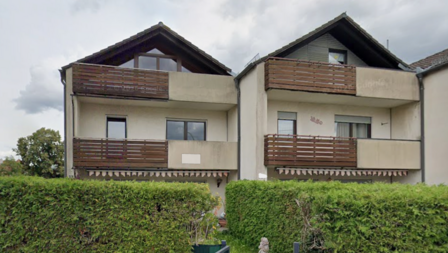 Wohngebäude mit Holzbalkonen und cremefarbener Fassade, davor hohe Hecke