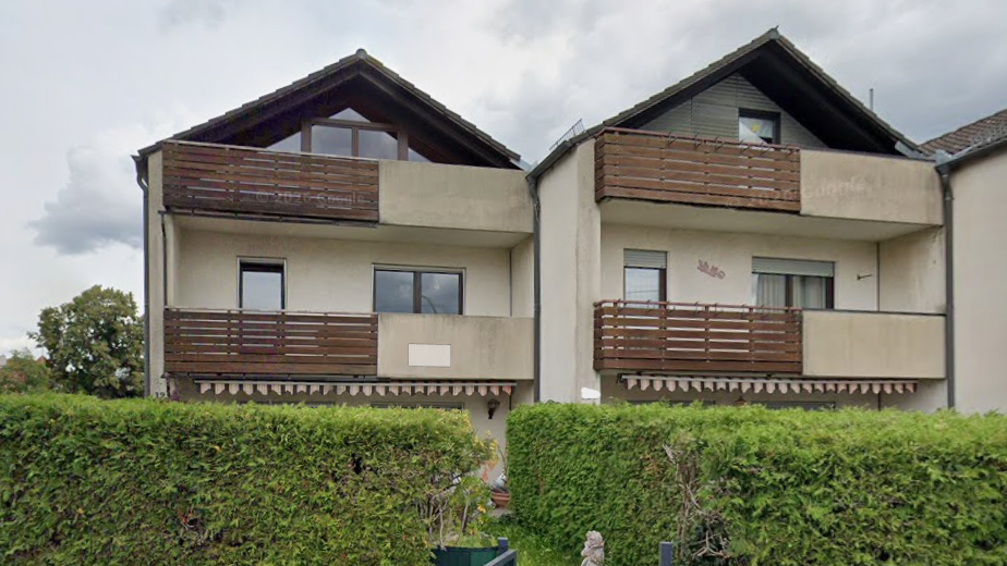 Wohngebäude mit Holzbalkonen und cremefarbener Fassade, davor hohe Hecke