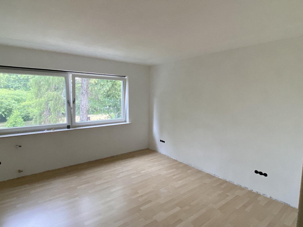 Leerer Raum mit weißer Wand, großem Fenster und hellem Laminatboden.