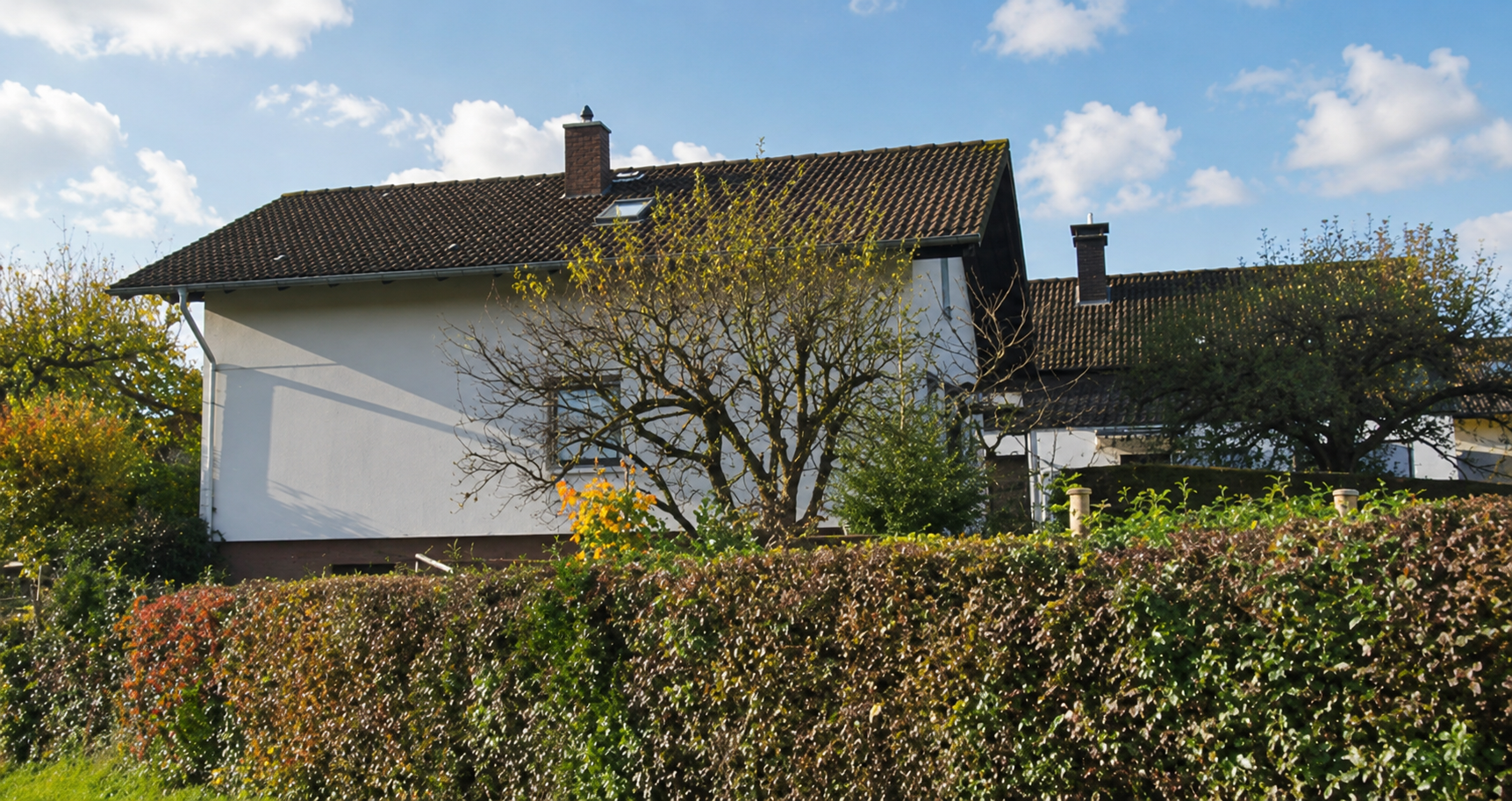 Weißes Wohnhaus mit Ziegeldach hinter Hecke und Baum, blauer Himmel mit Wolken