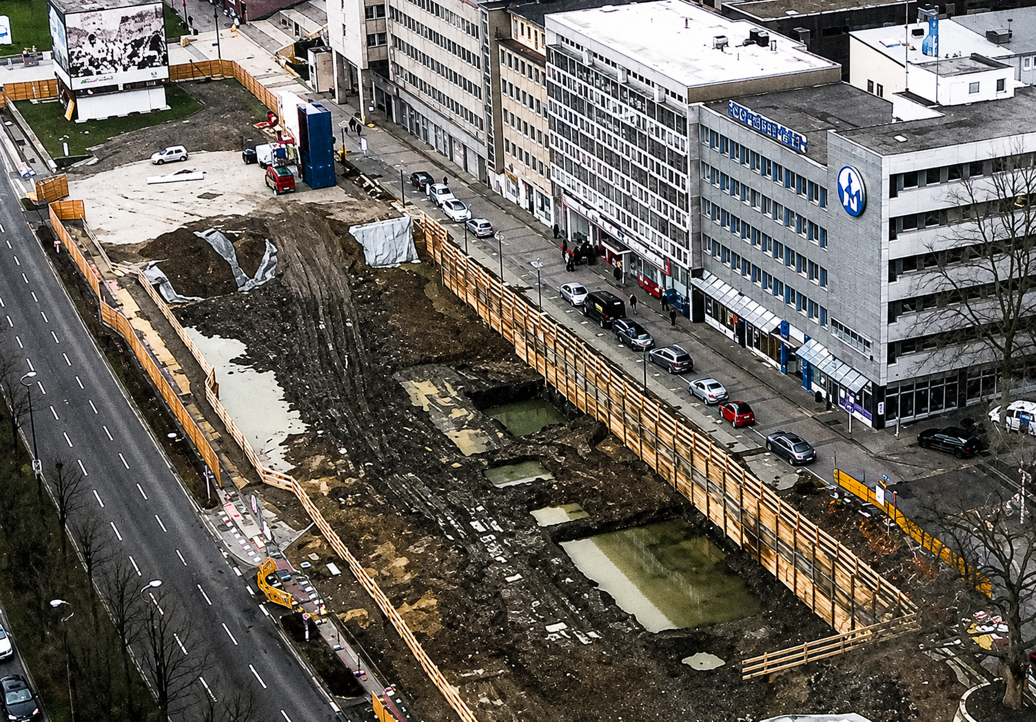 Luftaufnahme von Baugrube mit Erdwall, Holzabsperrung und mehreren Pfützen neben Straße