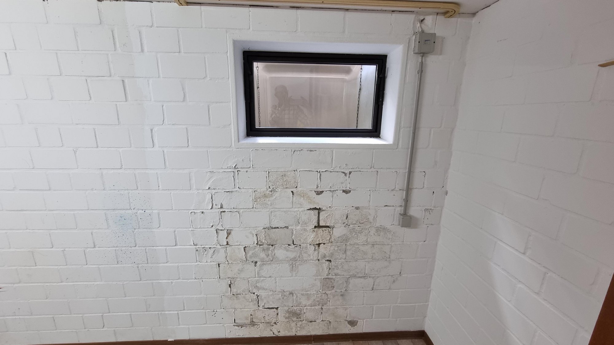 Weiß gestrichene Ziegelwand mit abgeplatzter Farbe und grauen Flecken unter einem kleinen Fenster.