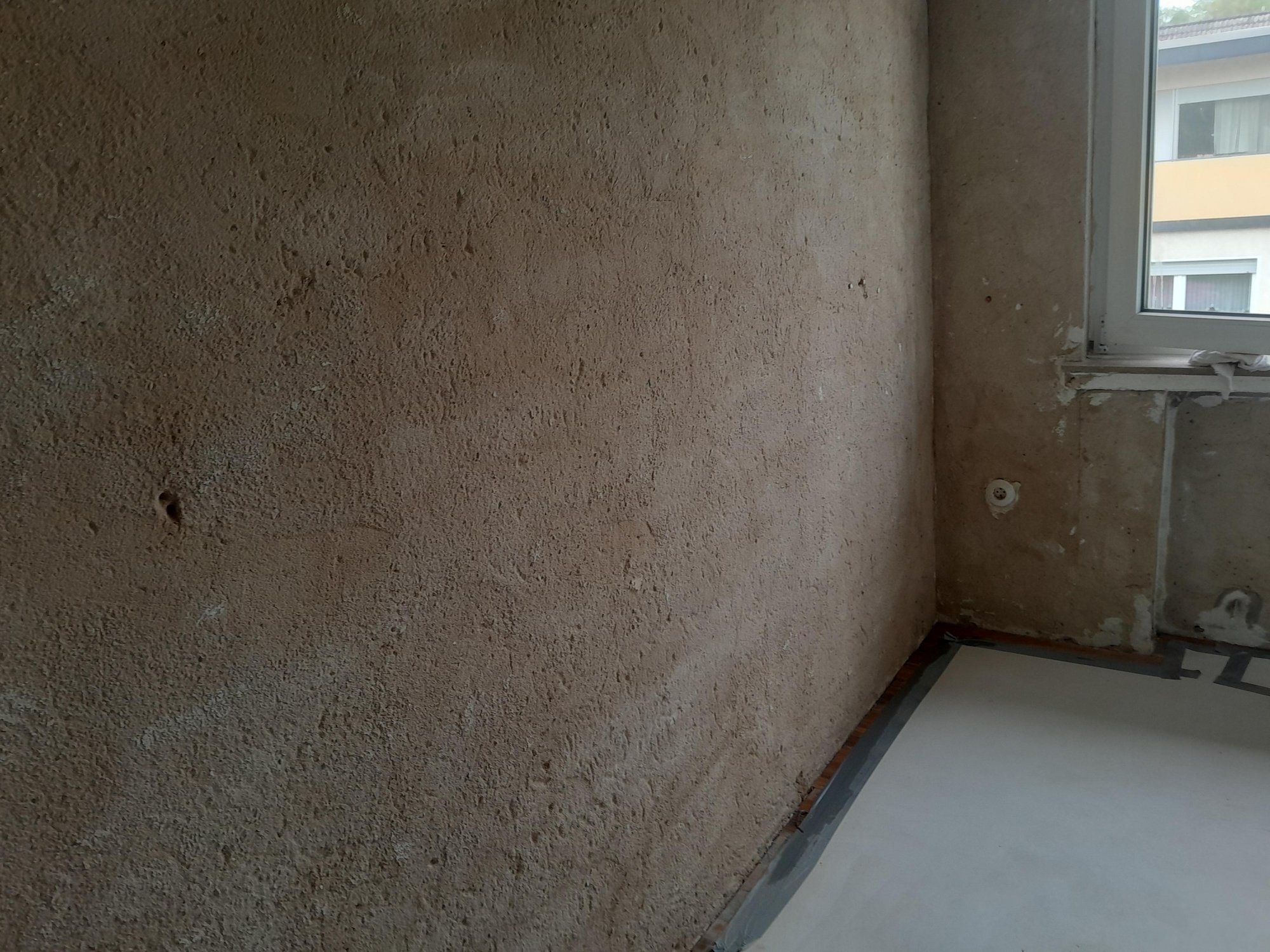 Raue beige Wandoberflaeche im Zimmer neben Fenster, Boden mit Abdeckfolie.