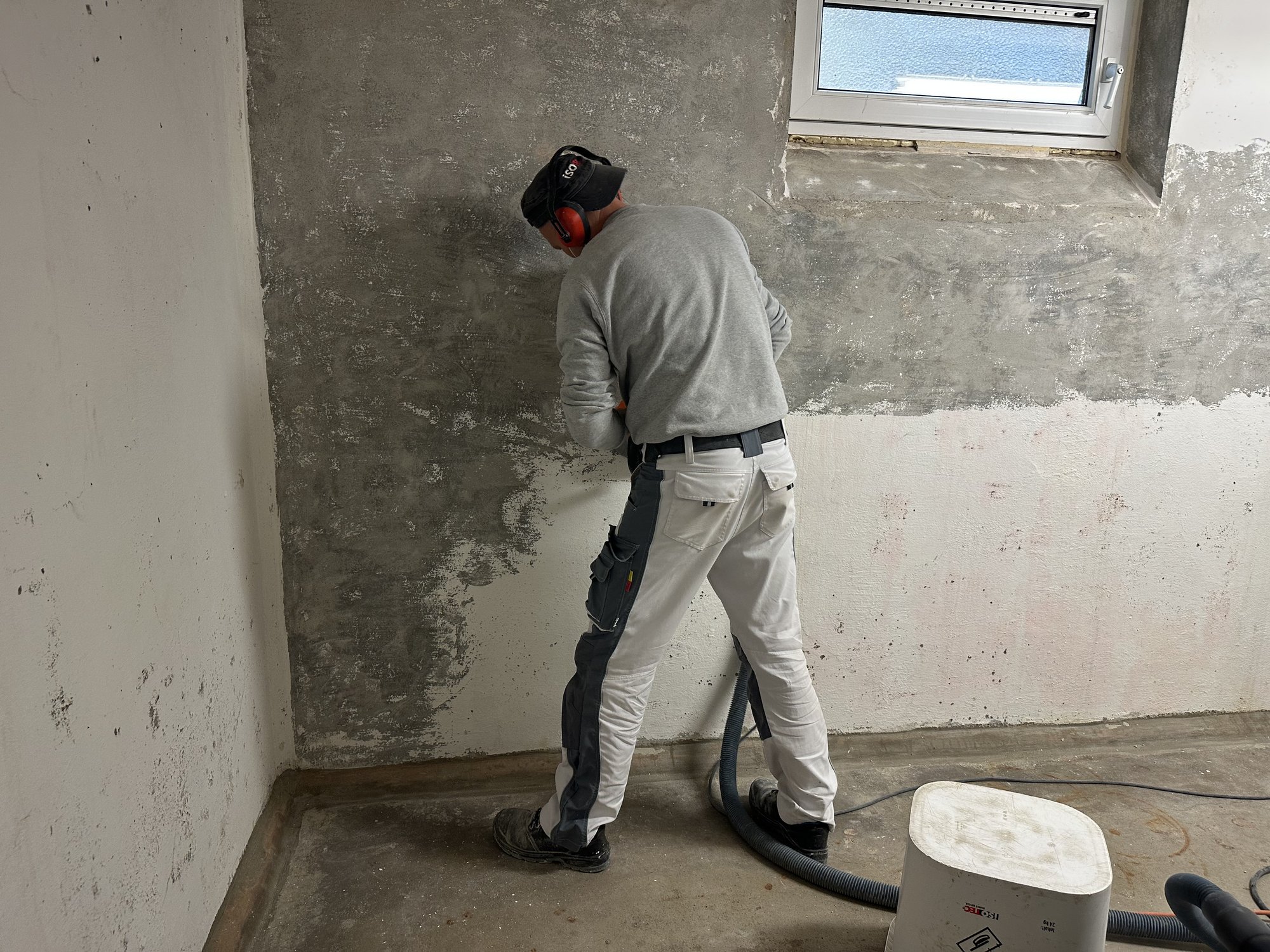 Arbeiter schleift graue Masse an Kellerwand neben Fenster ab.