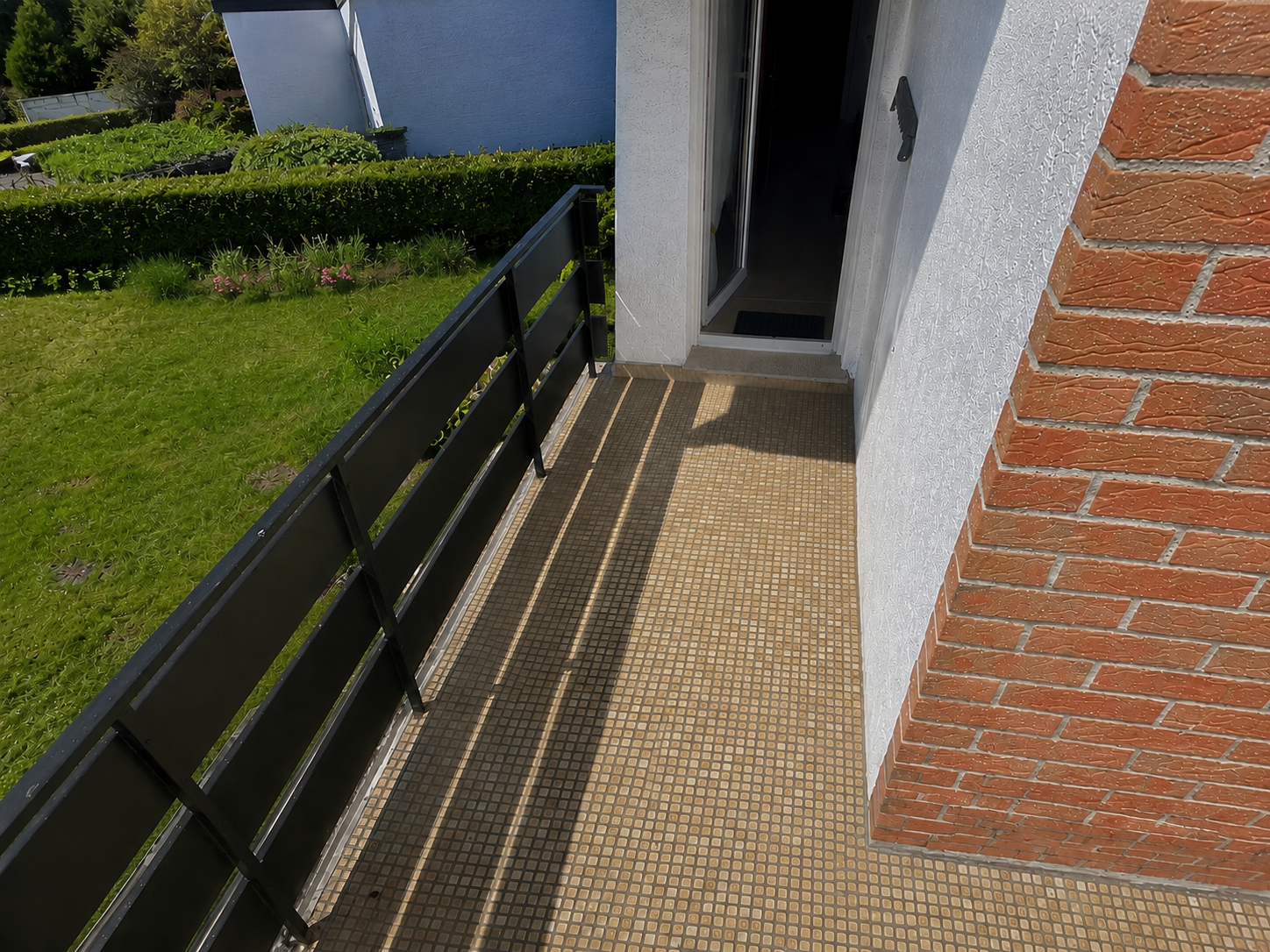Schmaler Balkon mit Fliesenboden, schwarzem Geländer und offener Tür am Ziegelpfeiler.