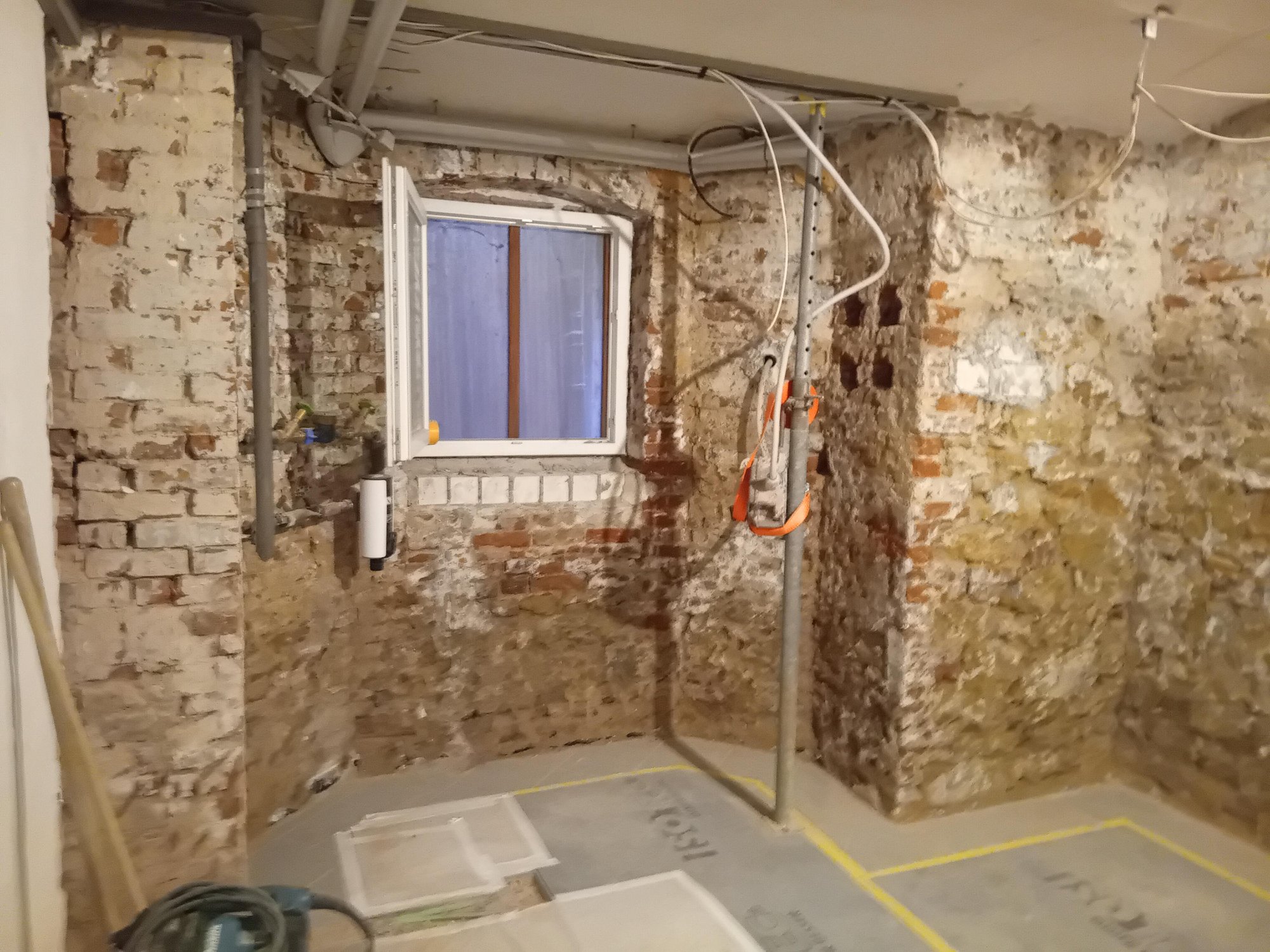 Freigelegte Ziegelwand mit Fenster, Rohren und Kabeln in renoviertem Kellerraum