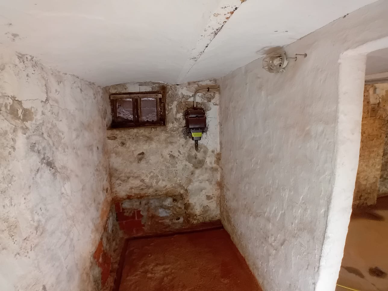 Feuchter Kellerraum mit abblätterndem Putz, kleinem Fenster und Stromkasten an der Wand