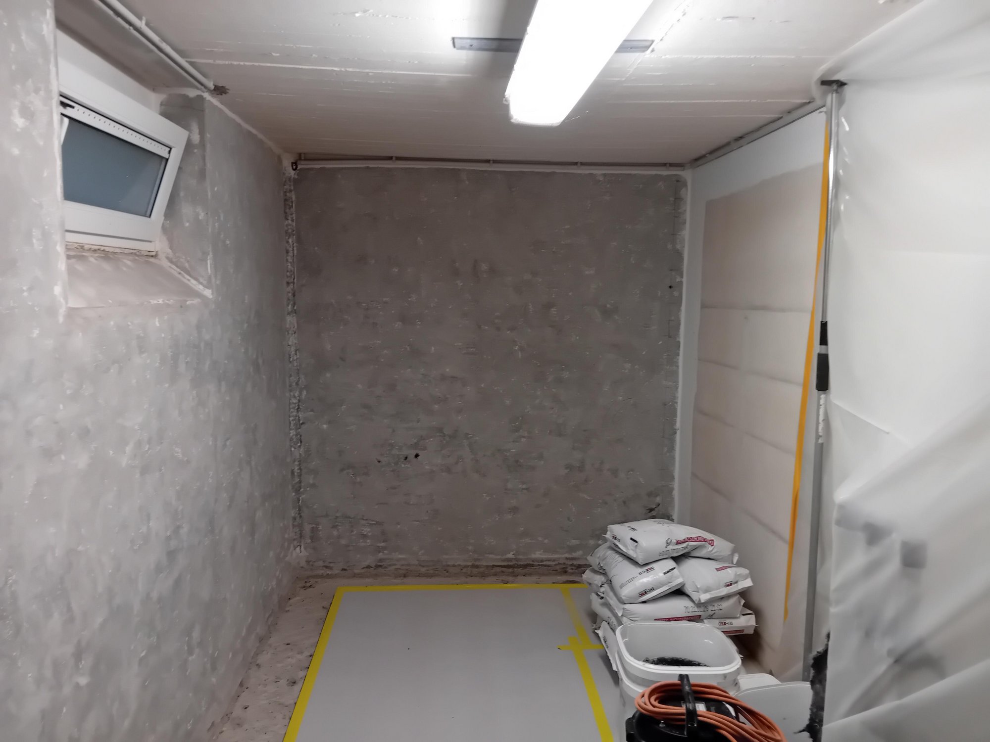 Leerer Kellerraum mit grauer Betonwand, Baustoffsäcken und Baustellenleuchte an der Decke