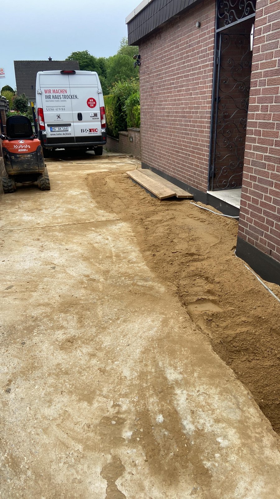 ISOTEC Transporter vor Backsteinhaus, Sand entlang Fundamentgraben neben Einfahrt.