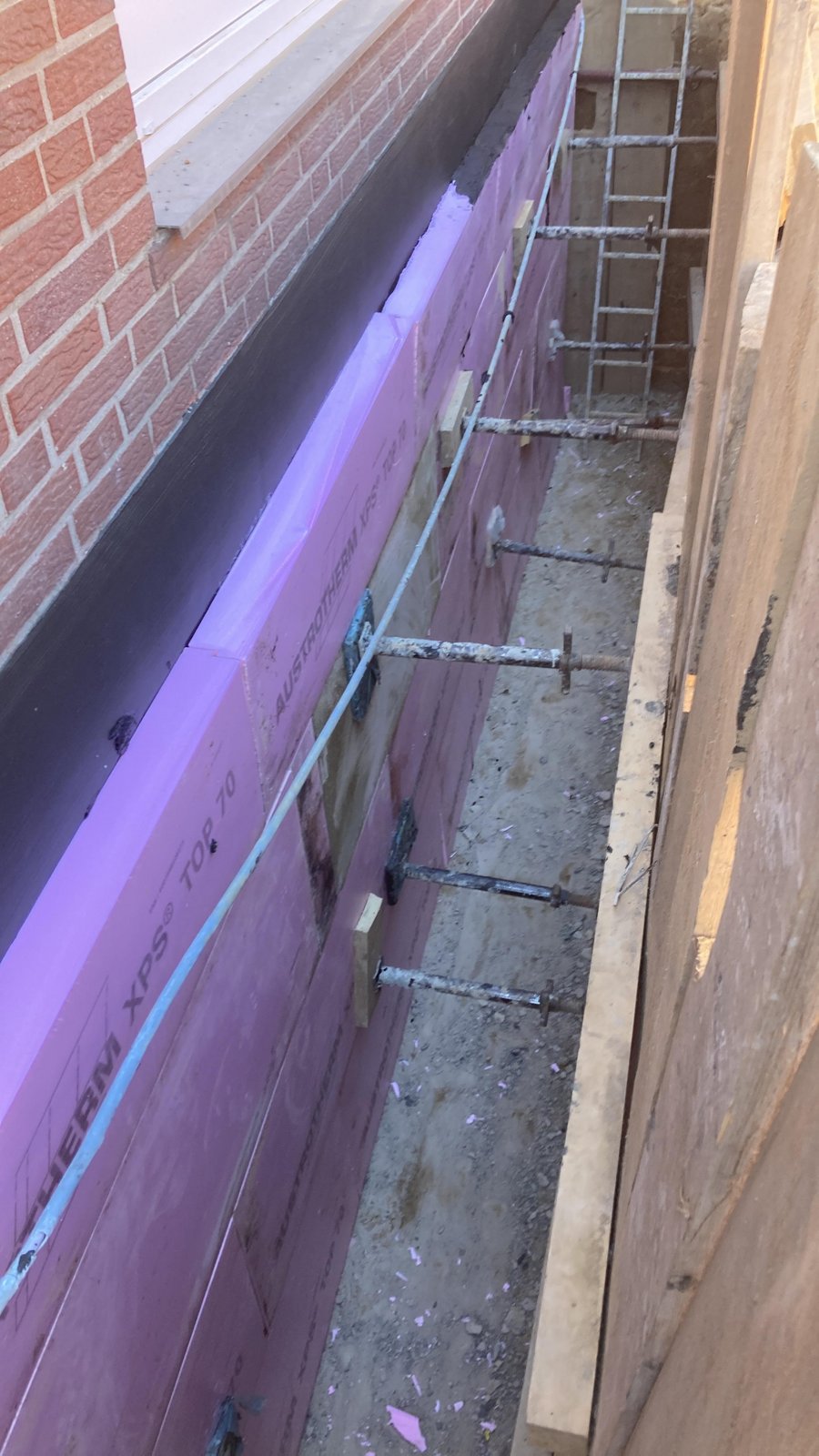 Schmaler Baugraben mit rosa Dämmplatten, Gerüststangen und Leiter neben Ziegelwand.