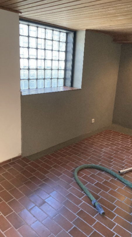 Raum mit Glasbausteinfenster, graue Wand und Terrakottafliesen, grüner Schlauch liegt auf dem Boden.