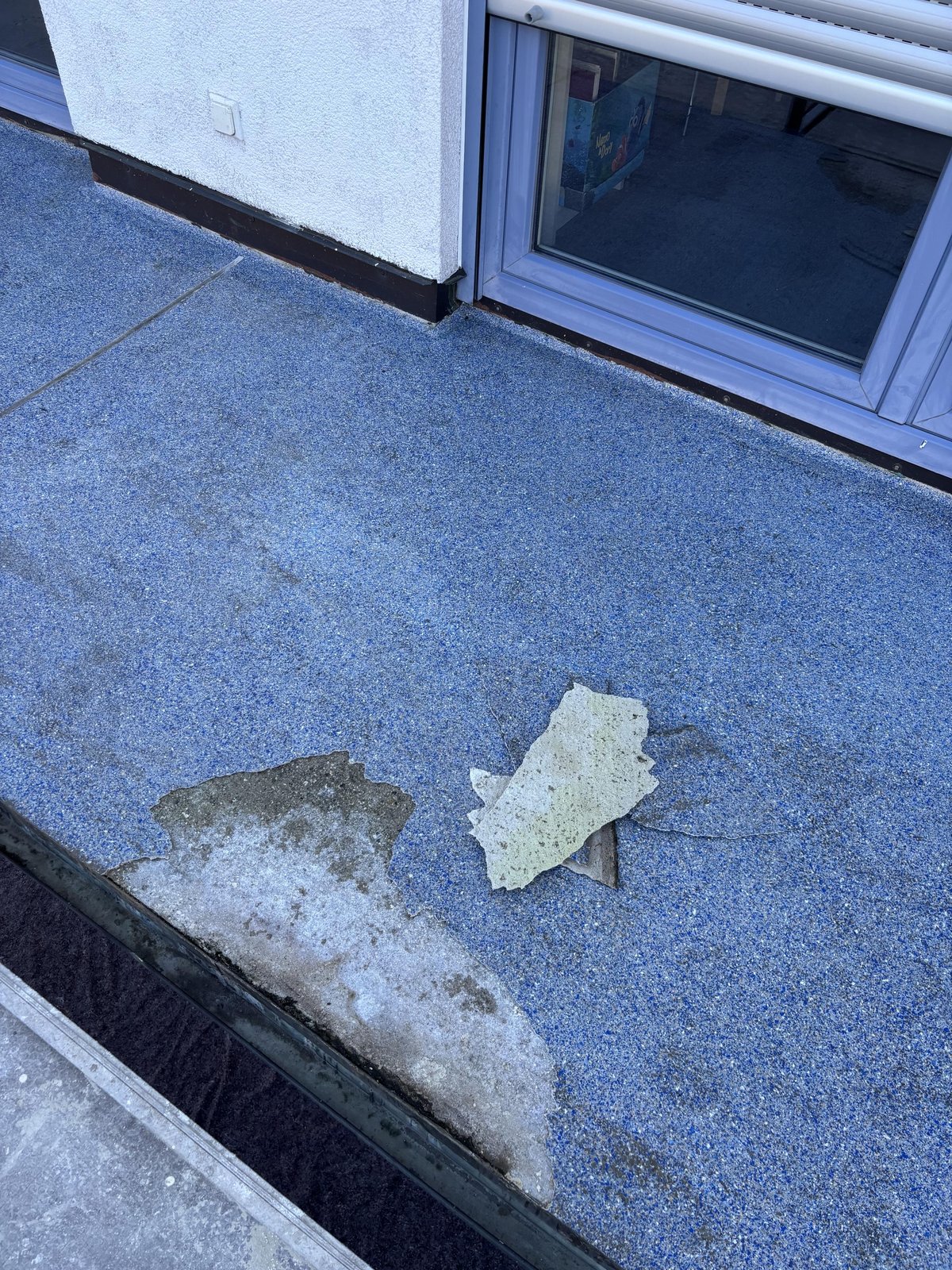 Abgelöste blaue Bodenbeschichtung auf Balkon, helle Platte liegt auf Schadstelle.