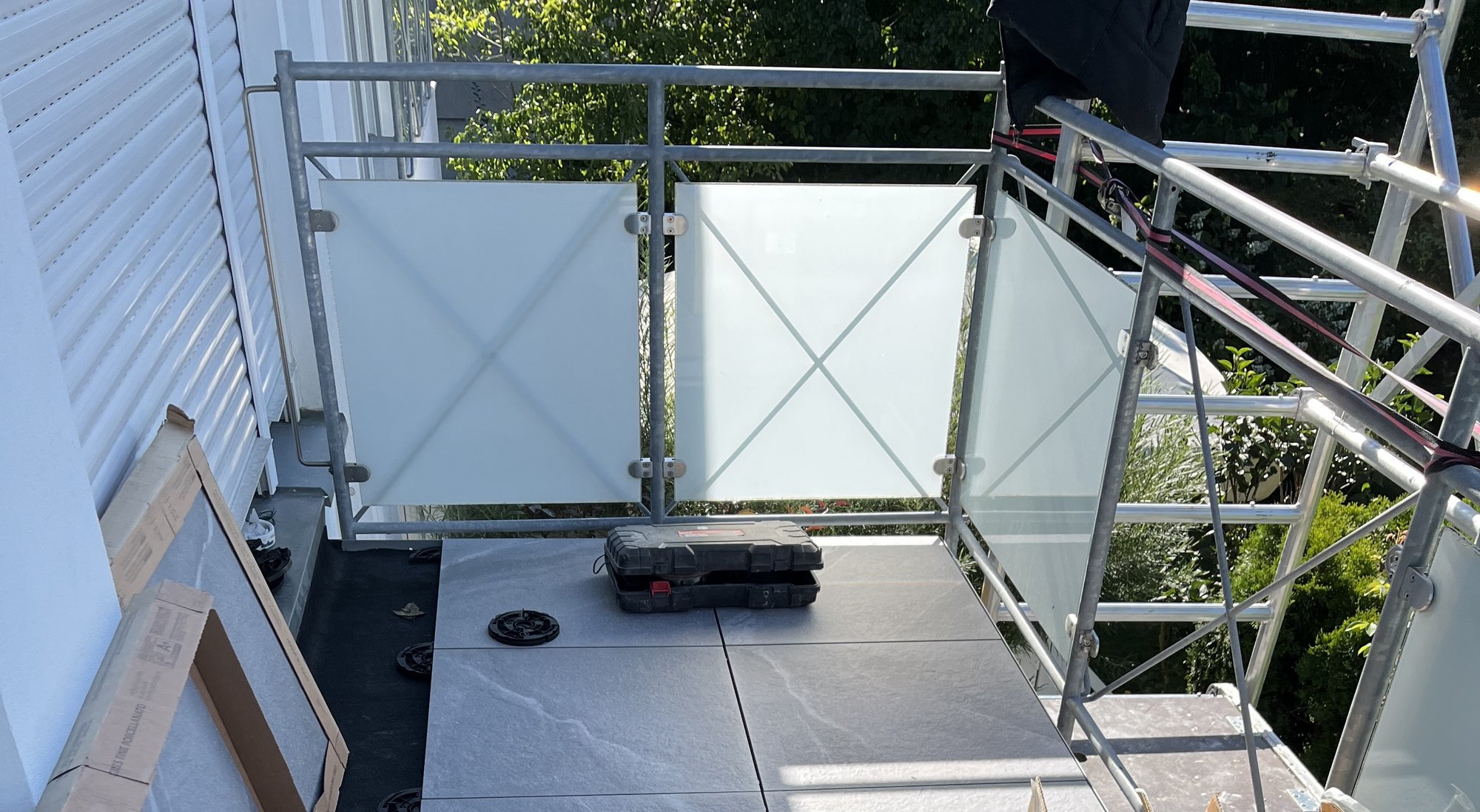 Geruest steht vor Balkon mit grauen Bodenplatten und Glasgelaender.