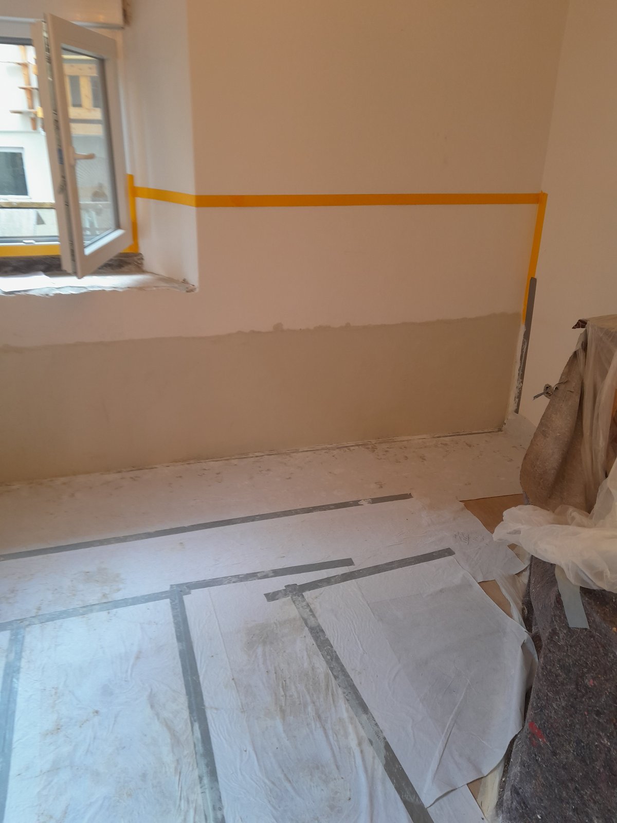 Abgeklebte Zimmerwand mit aufgetragener heller Masse im unteren Bereich.