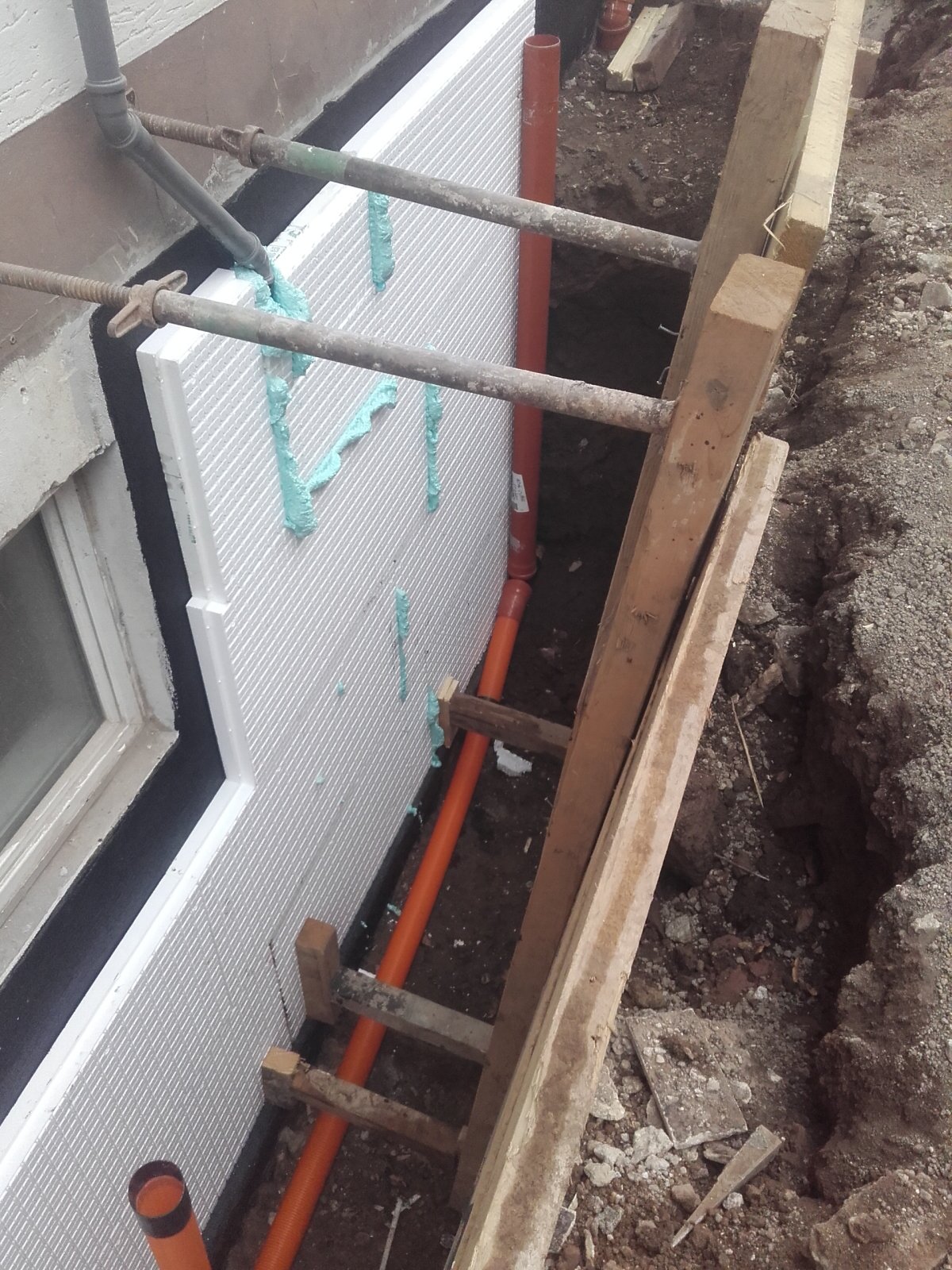 Weiße Dämmplatten mit grünem Schaum an Kellerwand neben orange Leitungen im Graben