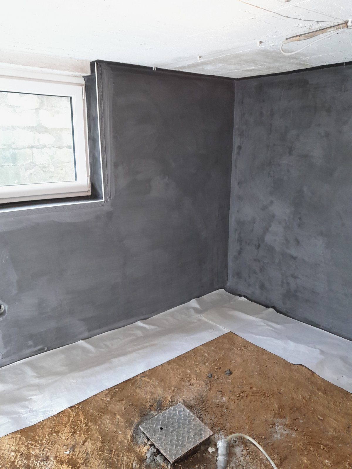 Mit dunkler Masse beschichtete Kellerwand mit offenem Bodenbereich