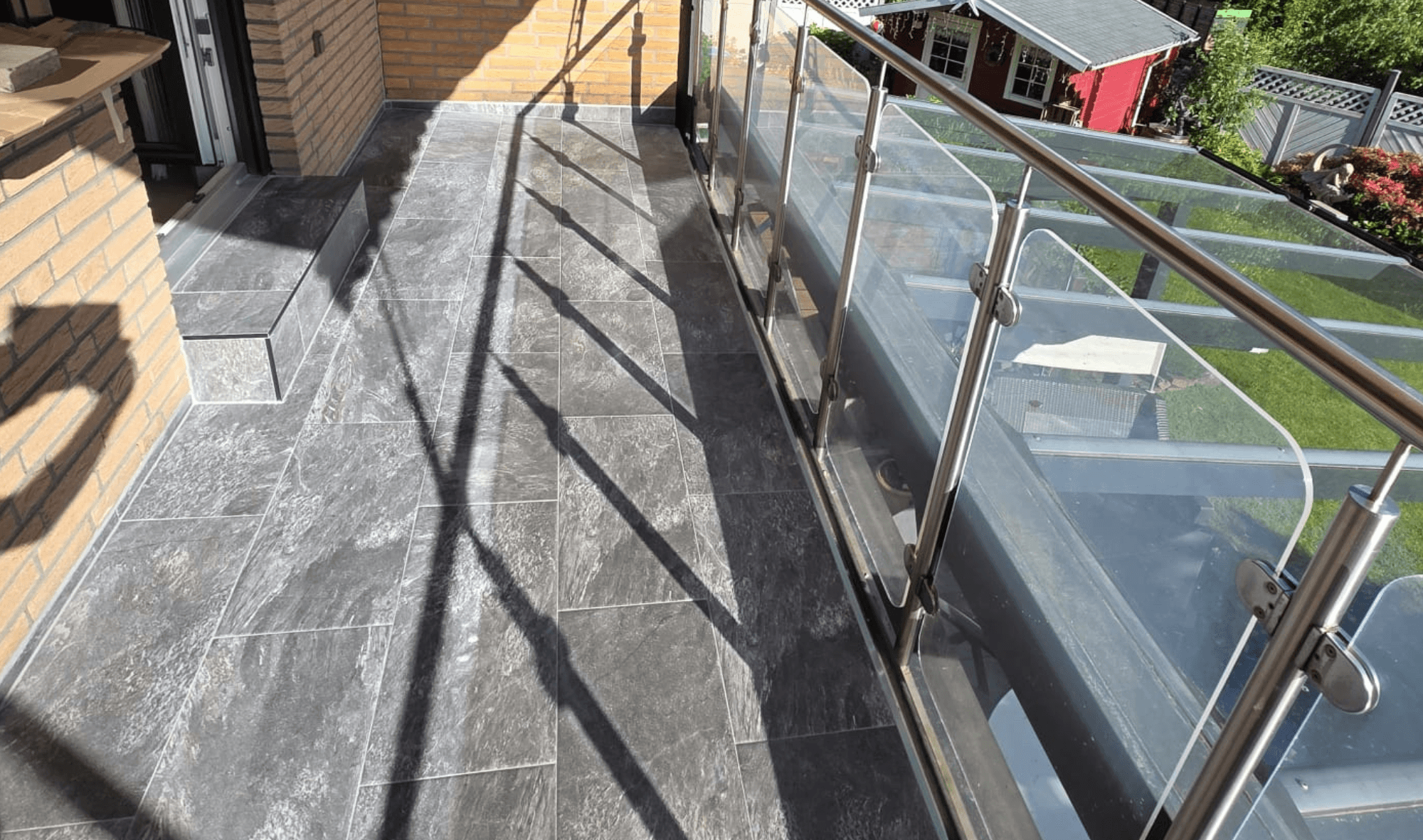 Balkon mit dunkelgrauen Bodenfliesen und Glasgeländer, Sonnenlicht wirft harte Schatten.