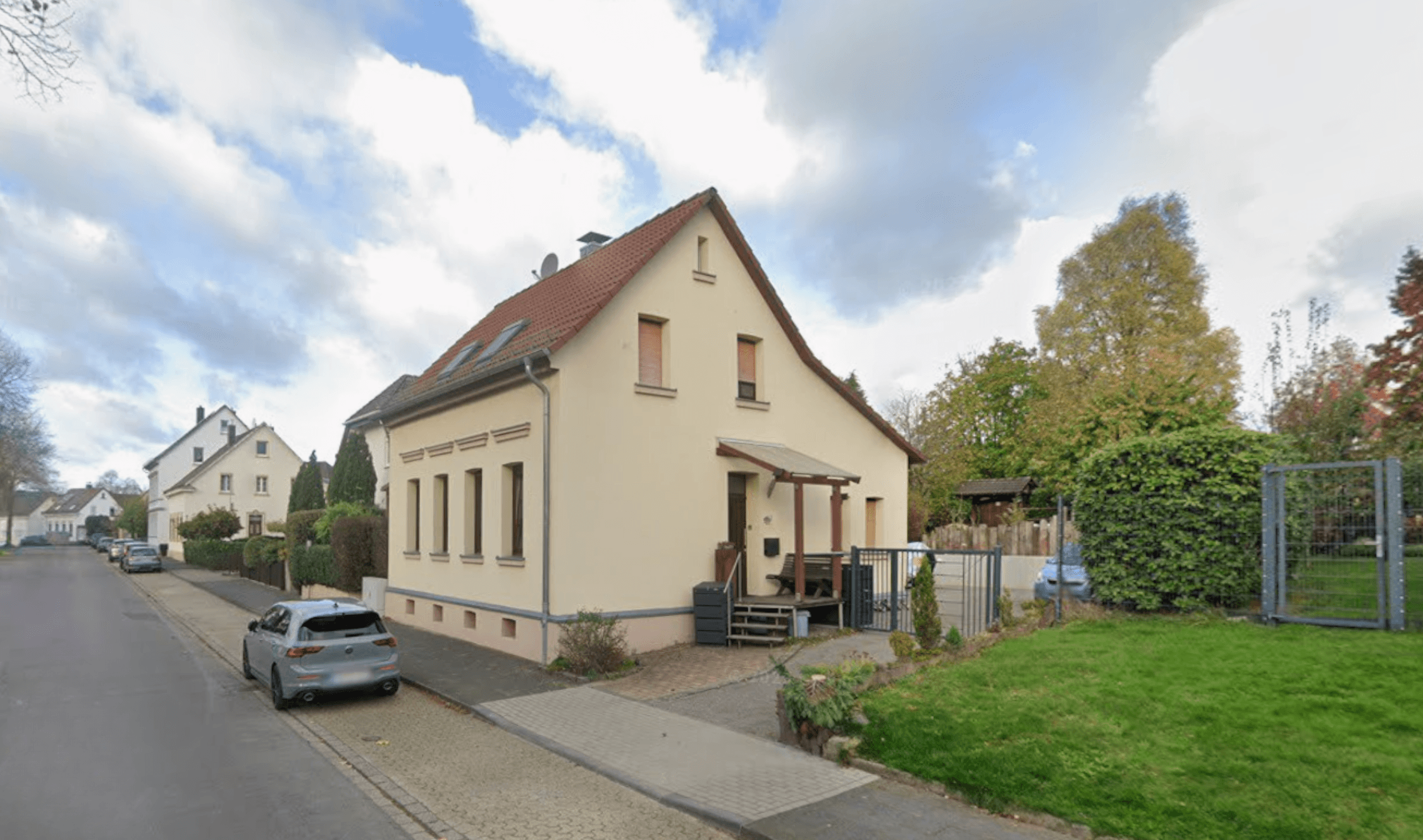 Beige Wohnhaus mit rotem Satteldach an Straße, Vorgarten und Parkplatz.