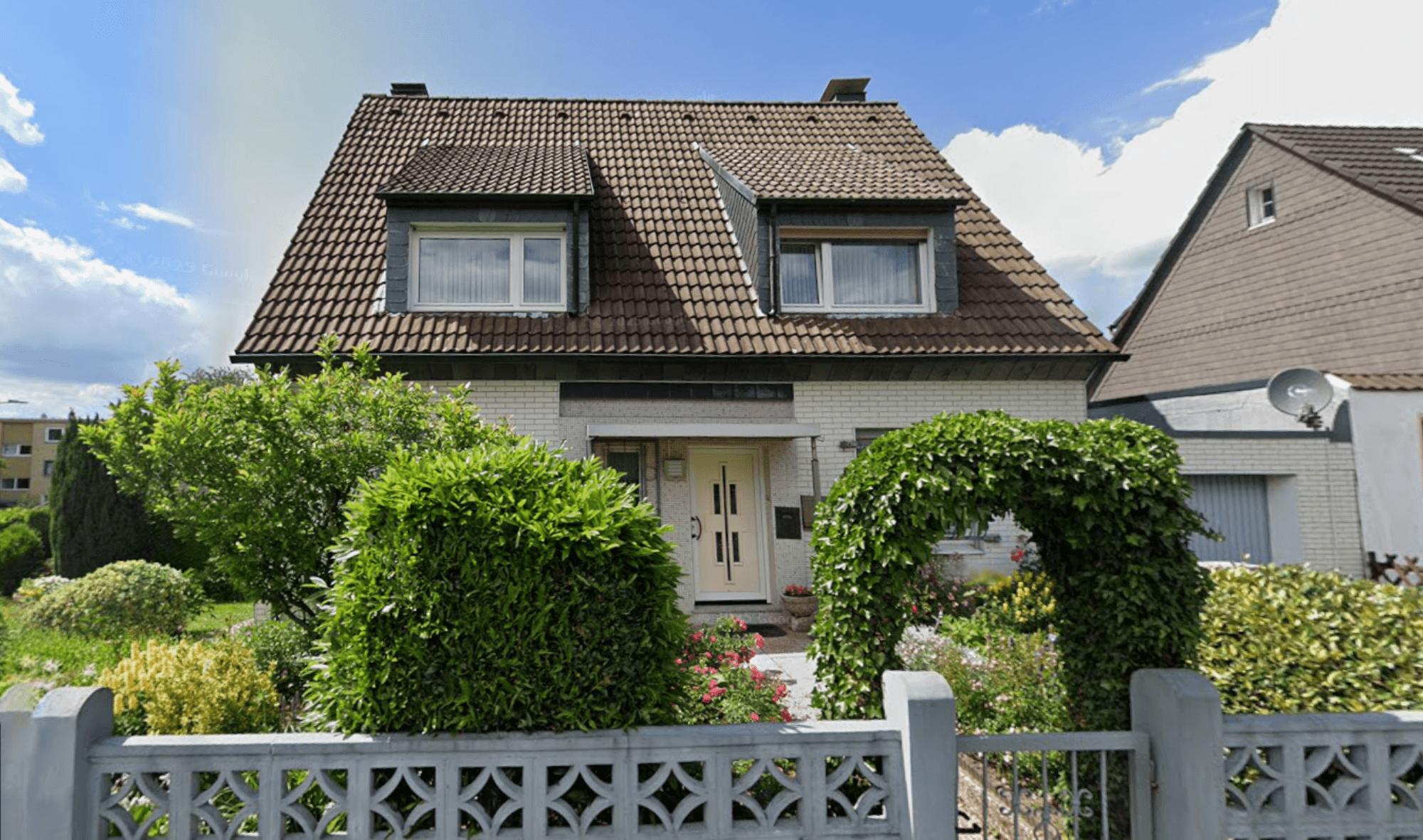 Einfamilienhaus mit braunem Ziegeldach, Vorgarten mit Heckenbogen und Zaun