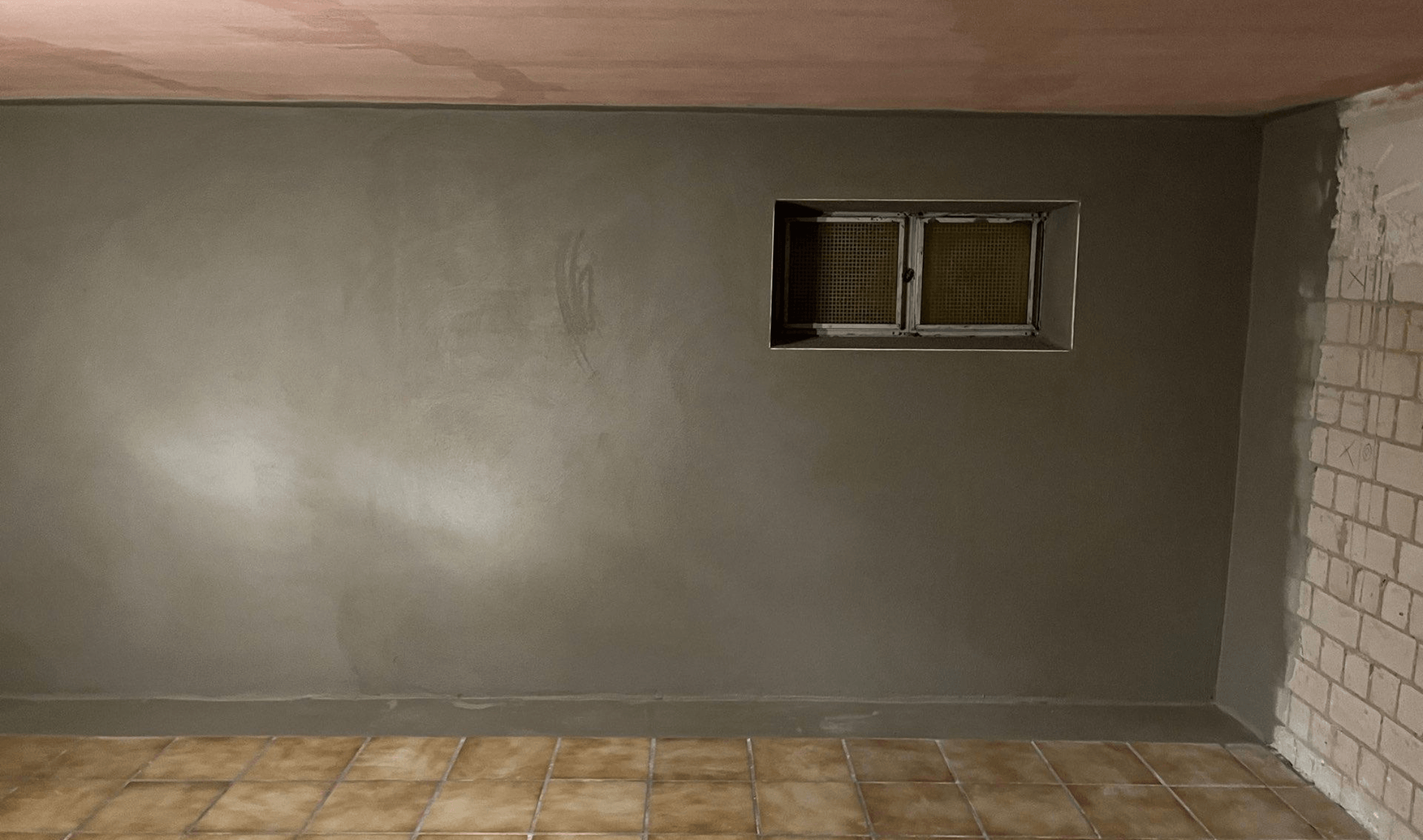 Graue Wand mit kleinem Kellerfenster und hellem Fleck, Innenraum mit Fliesenboden