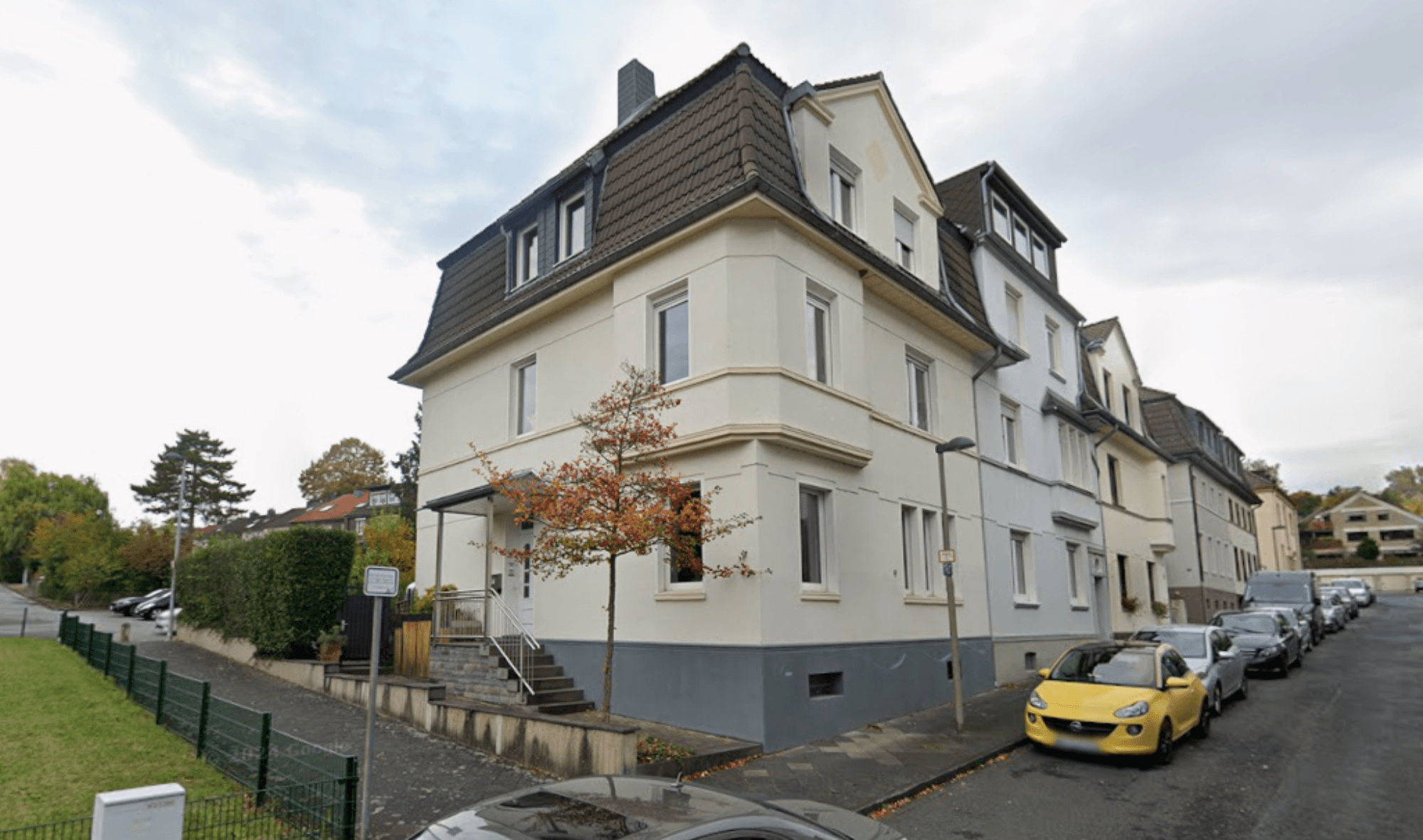 Mehrstöckiges helles Wohngebäude an Straße, parkende Autos, Herbstbaum im Vordergrund