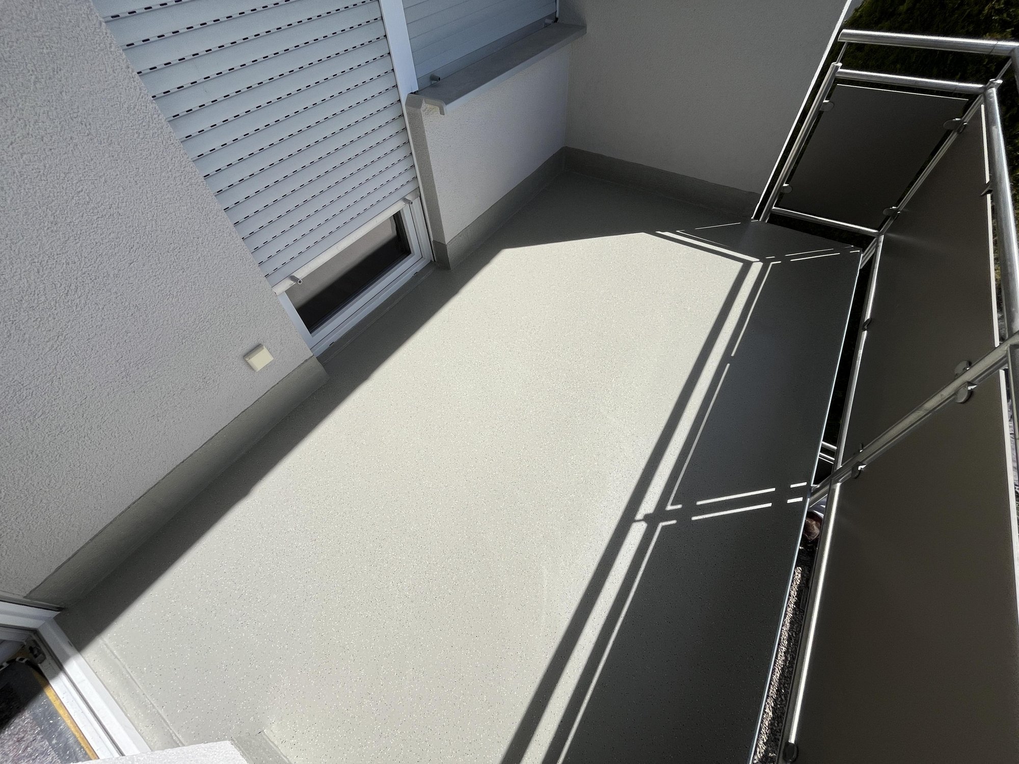 Sonniger Balkon mit grauem Bodenbelag, Metallgeländer und Fensterrollladen