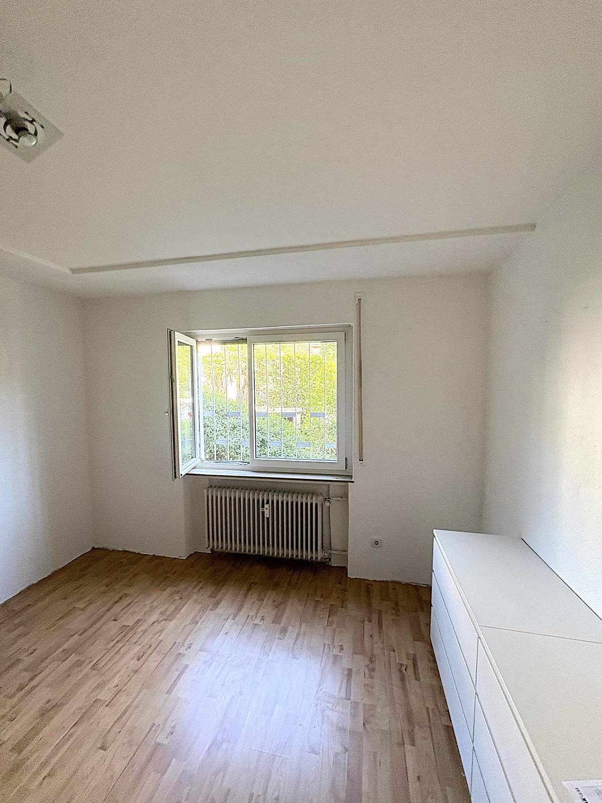 Leeres Zimmer mit Holzlaminat, Fenster mit Gitter und Heizkörper darunter