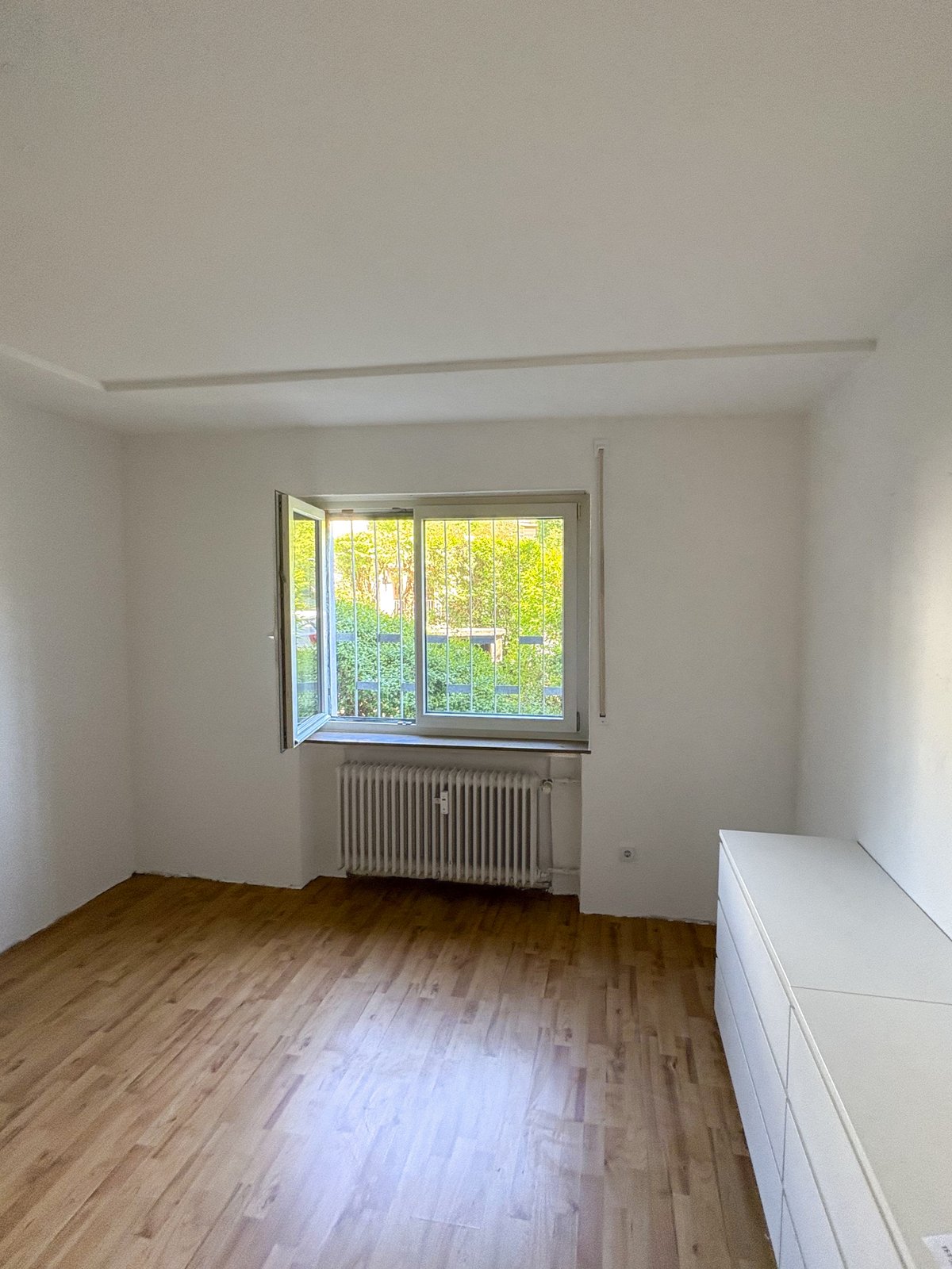 Leeres Zimmer mit Holzfußboden, geöffnetem Fenster mit Gitter und Heizkörper darunter