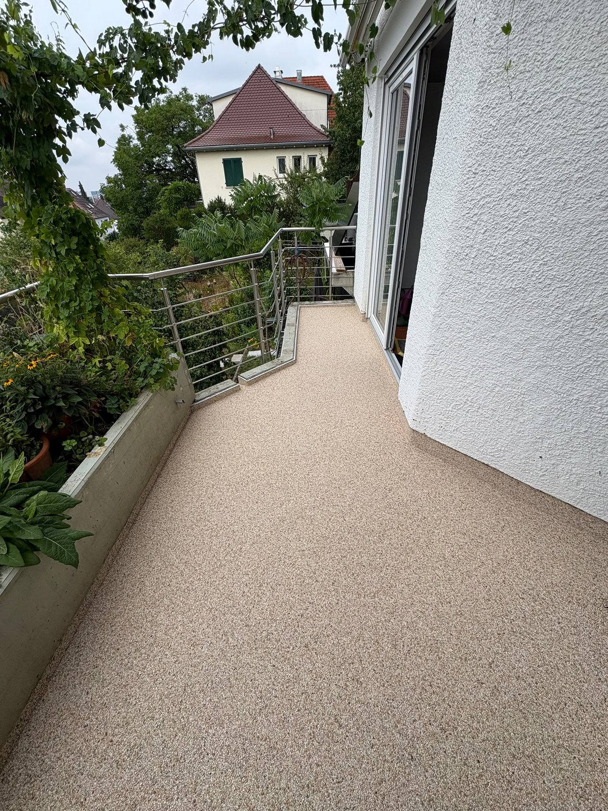 Balkon mit hellem Bodenbelag, Metallgeländer und Pflanzen, Blick auf Nachbarhaus.