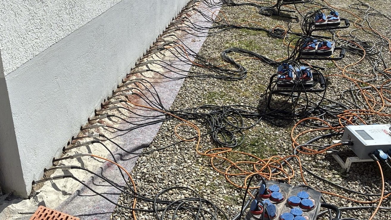 Kabel und Mehrfachsteckdosen liegen auf Kies neben verputzter Wand, viele Leitungen befestigt
