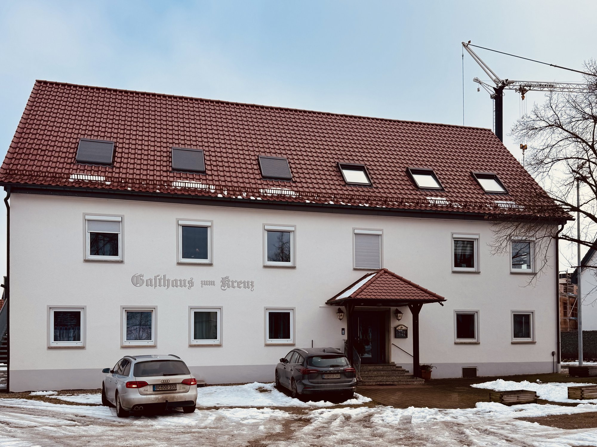 Weißes Gasthaus mit rotem Ziegeldach, Dachfenstern, parkenden Autos und Schnee
