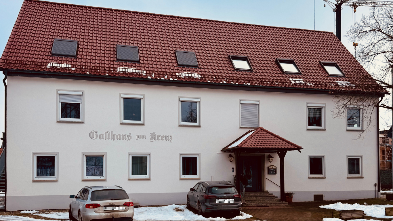 Weißes Gebäude mit rotem Ziegeldach, Schriftzug „Gasthaus zum Kreuz“ und zwei Autos davor.