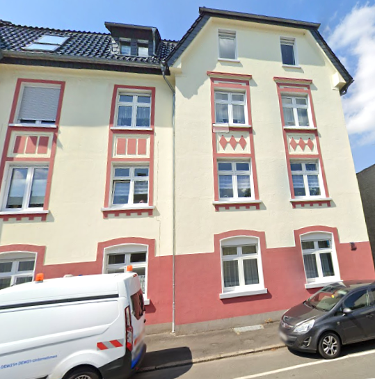 Mehrstöckiges Wohnhaus mit hellgelber Fassade, roten Fensterrahmen und Satteldach, Straßenansicht.