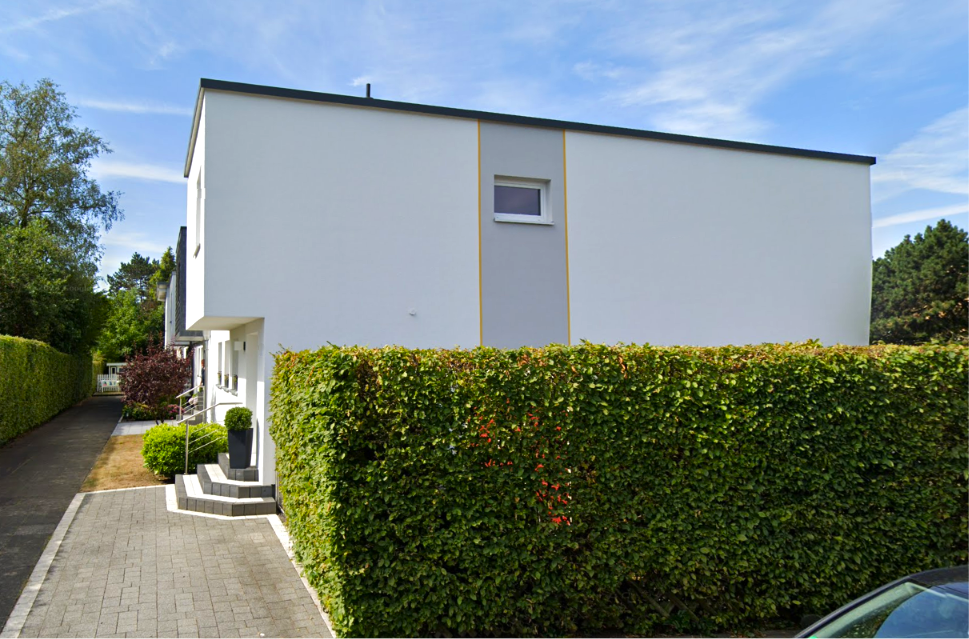 Weißes Flachdachhaus hinter hoher Hecke, seitlicher Weg und Eingang mit Treppe.