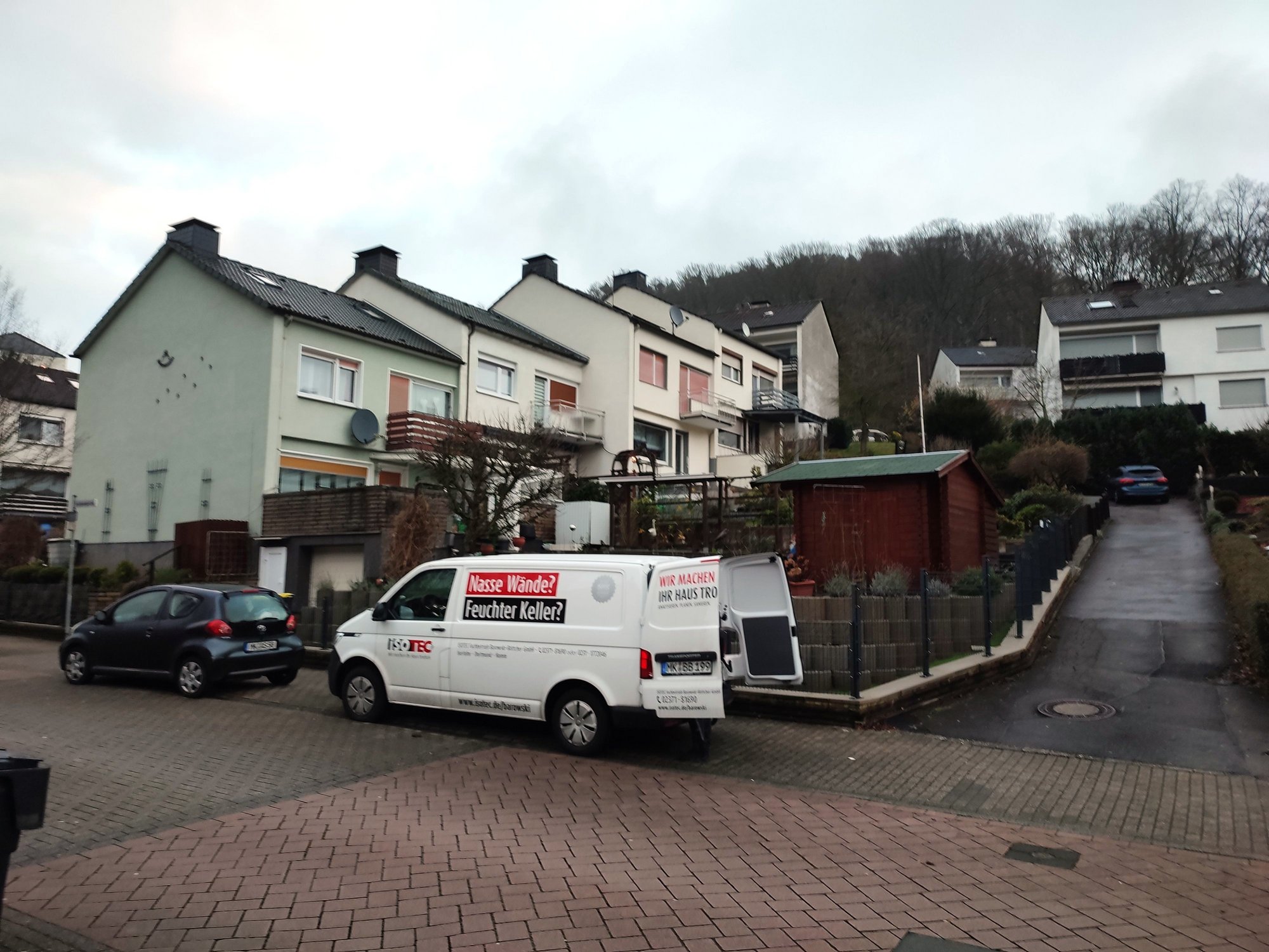 ISOTEC Transporter parkt vor Reihenhäusern in Wohnstraße, Hanglage mit Bäumen im Hintergrund.