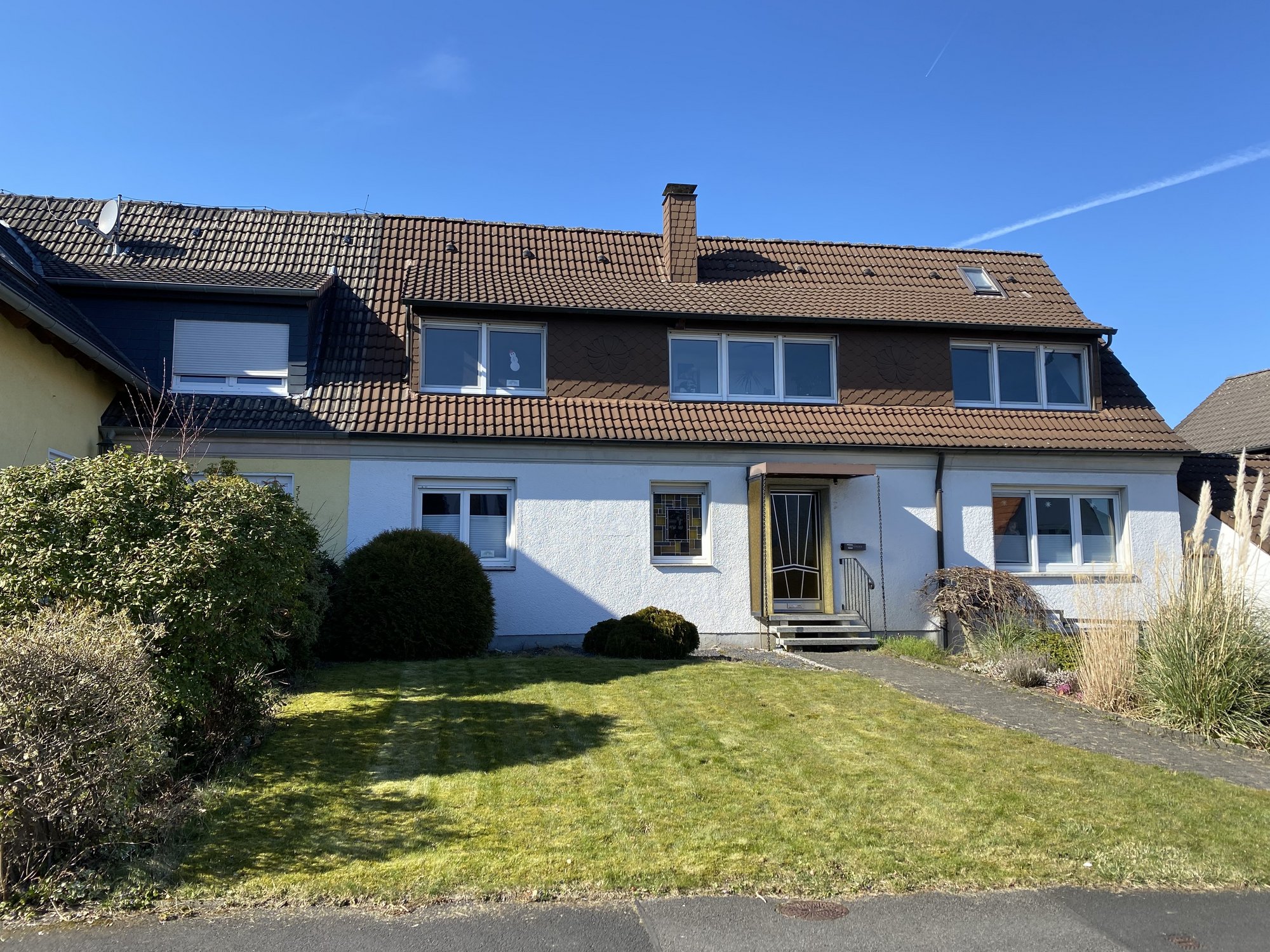 Einfamilienhaus mit braunem Ziegeldach, weißen Wänden und Vorgarten bei blauem Himmel