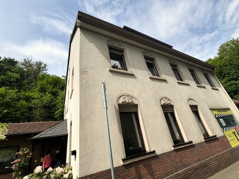Schrägansicht eines hell verputzten Hauses mit mehreren Fenstern und Ziegelsockel.