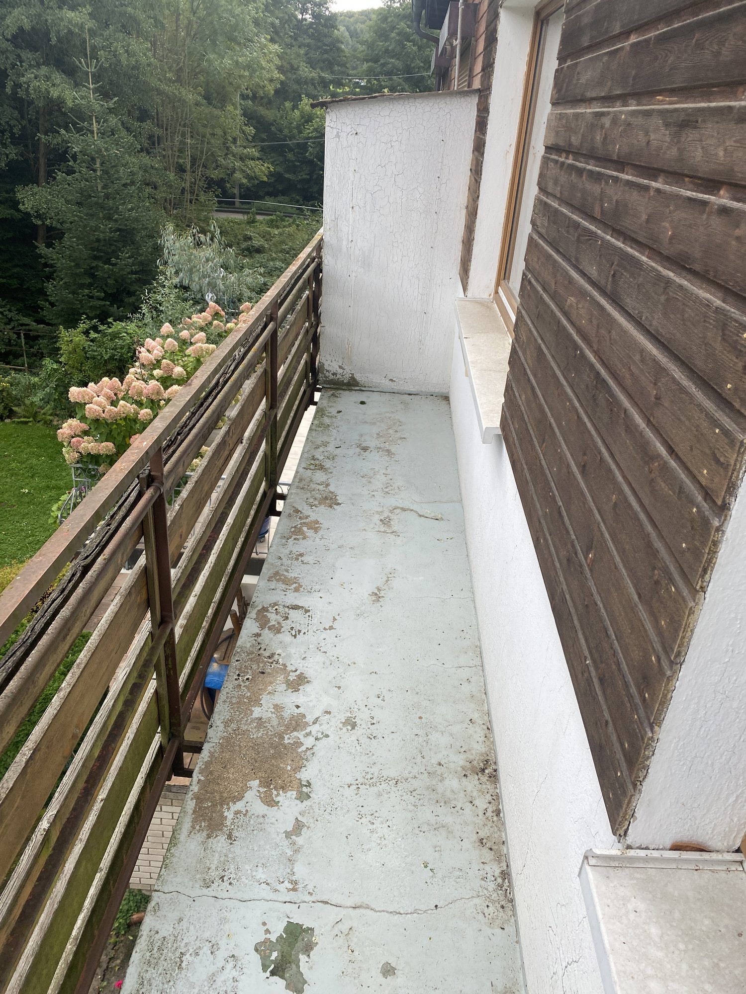 Schmaler Balkon mit rissigem, fleckigem Bodenbelag und verwittertem Holzgeländer.