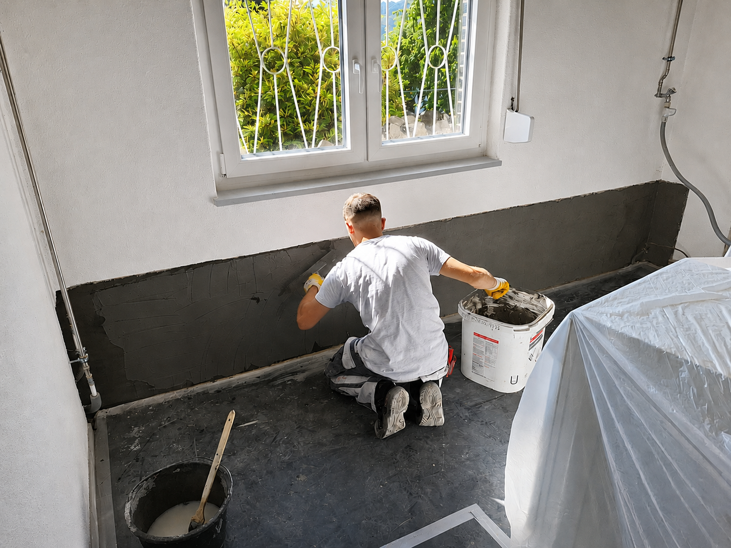 Arbeiter trägt graue Masse auf Wand im Zimmer unter Fenster auf.