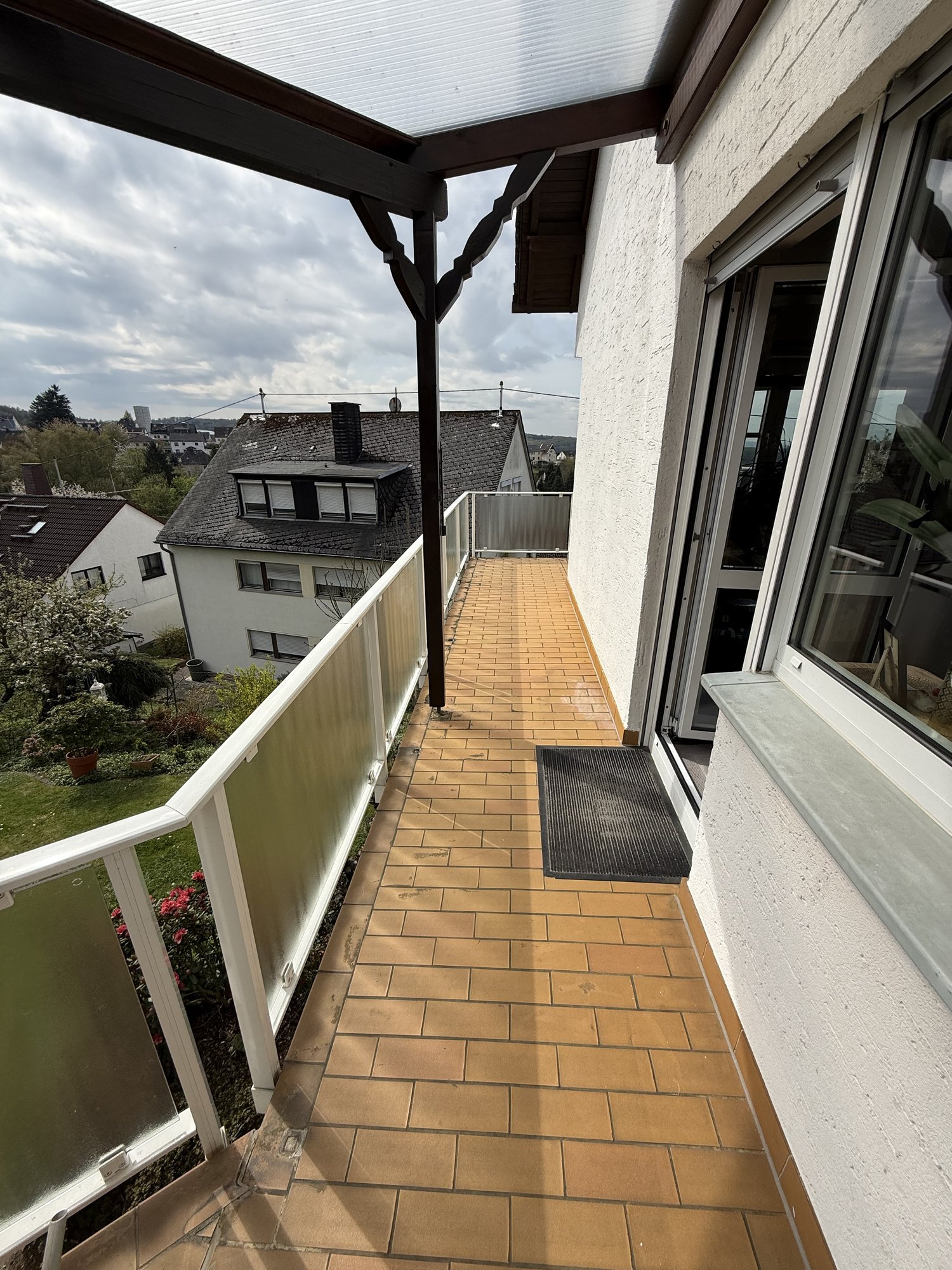 Schmaler Balkon mit braunen Fliesen, Glasgeländer und Blick auf Wohnhäuser.