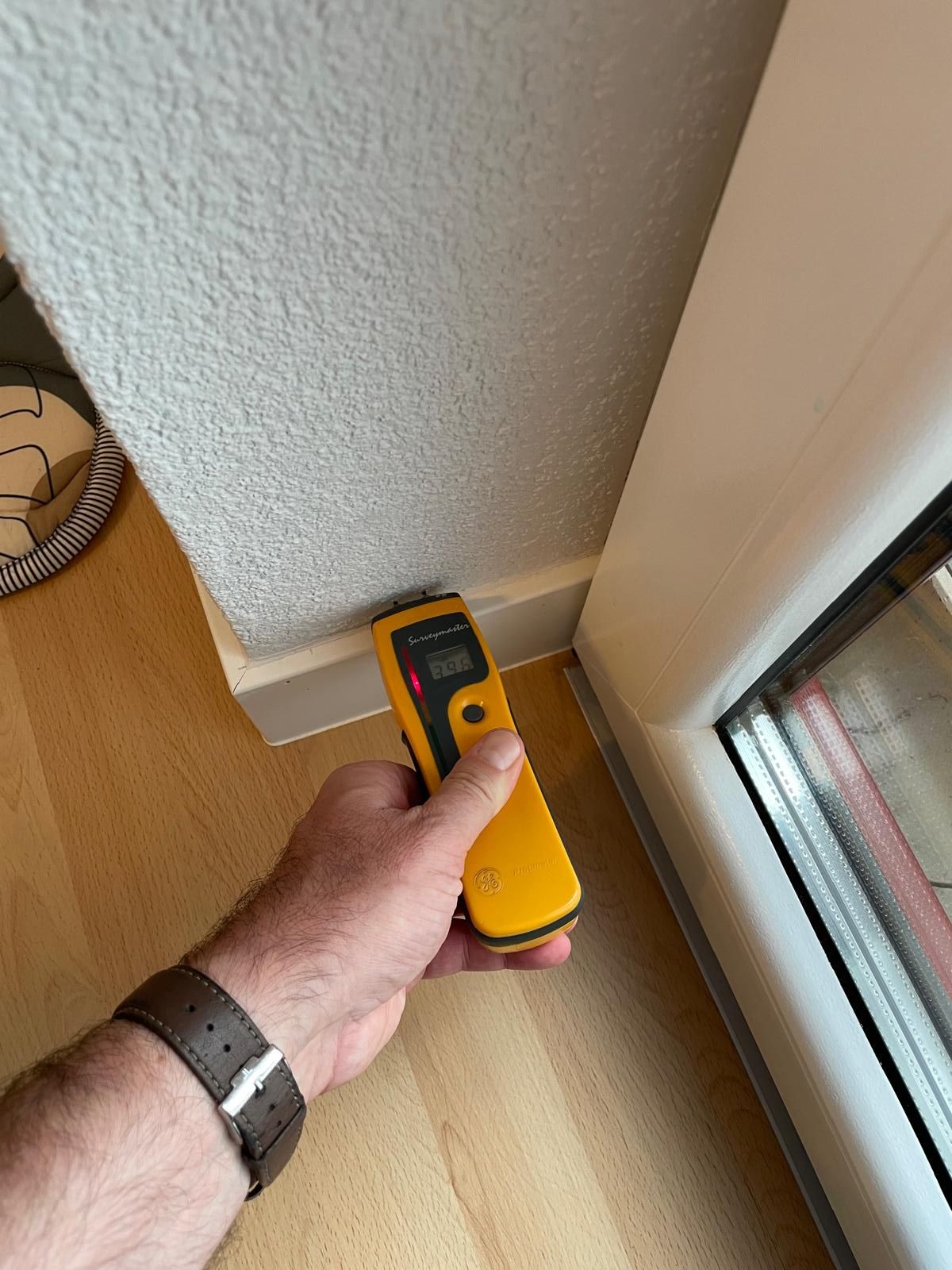 Hand hält Feuchtigkeitsmessgerät am Sockel neben Fensterrahmen.