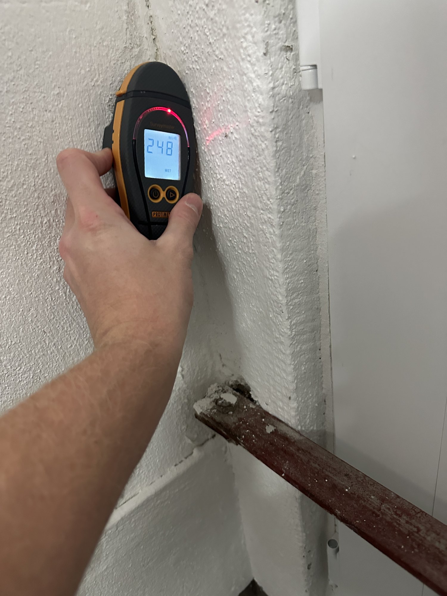 Hand hält Feuchtigkeitsmessgerät an strukturierte Wand, Display zeigt Messwert.