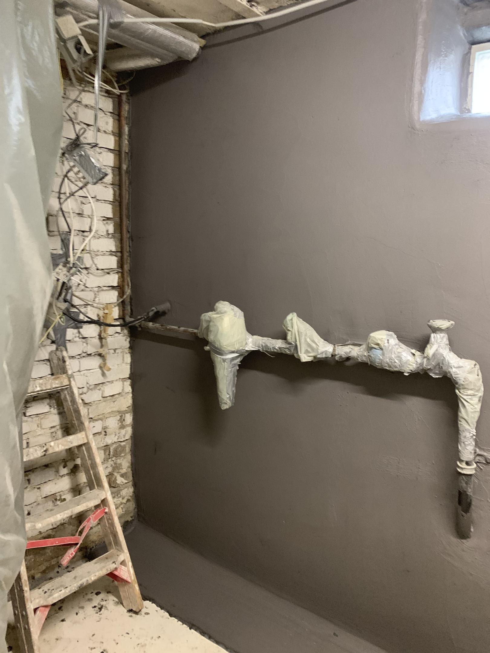 Graue Masse auf Kellerwand, Rohre mit Folie abgedeckt, freigelegte Ziegel und Leiter.