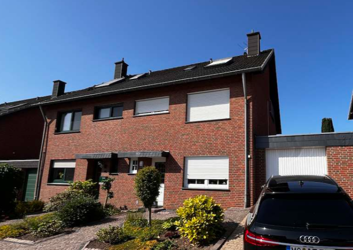 Zweigeschossiges Backsteinhaus mit Rolllaeden, Vorgarten und geparktem Auto vor Garage.