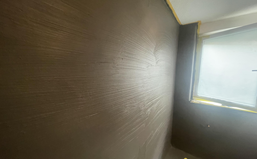 Schwarze Masse auf Wandfläche mit Spachtelstruktur neben abgeklebt­em Fenster.