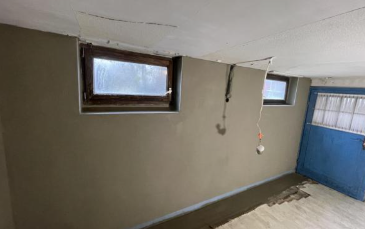 Kellerraum mit zwei kleinen Fenstern, beschädigter Decke und freihängendem Kabel