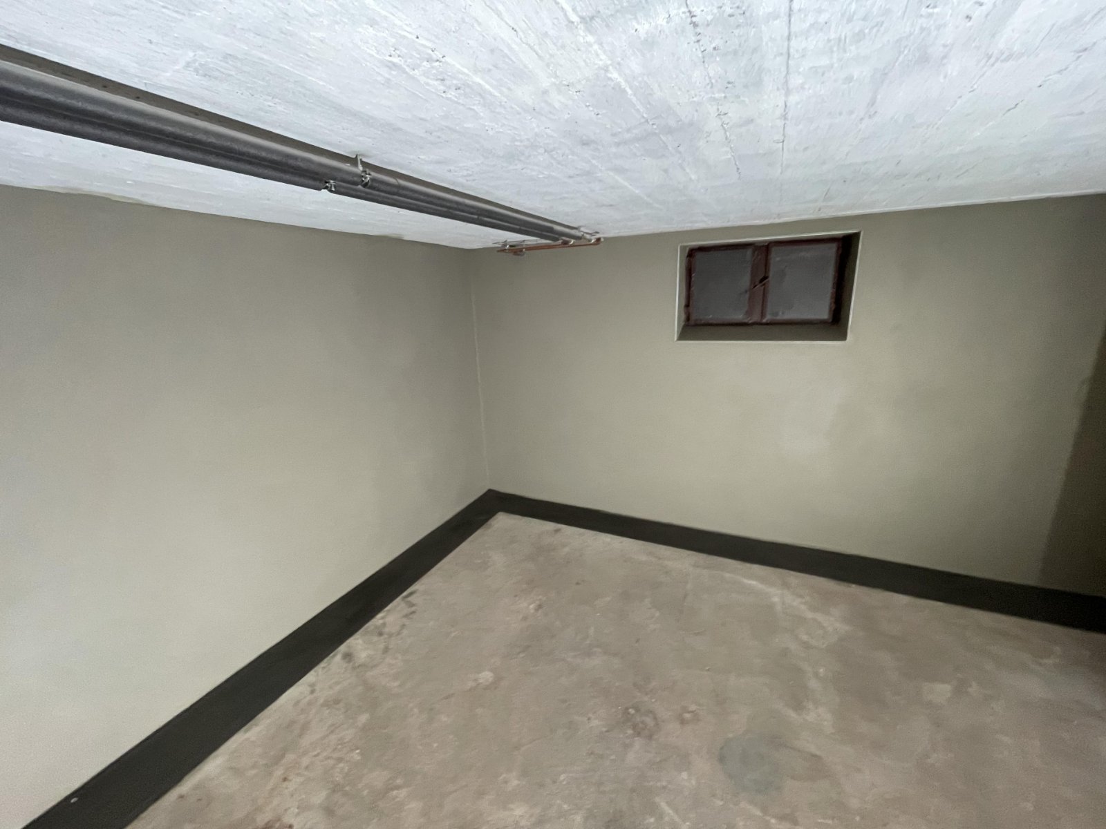 Leerer Kellerraum mit beige-grauen Wänden, Metallrohr an Betondecke und kleinem Fenster.